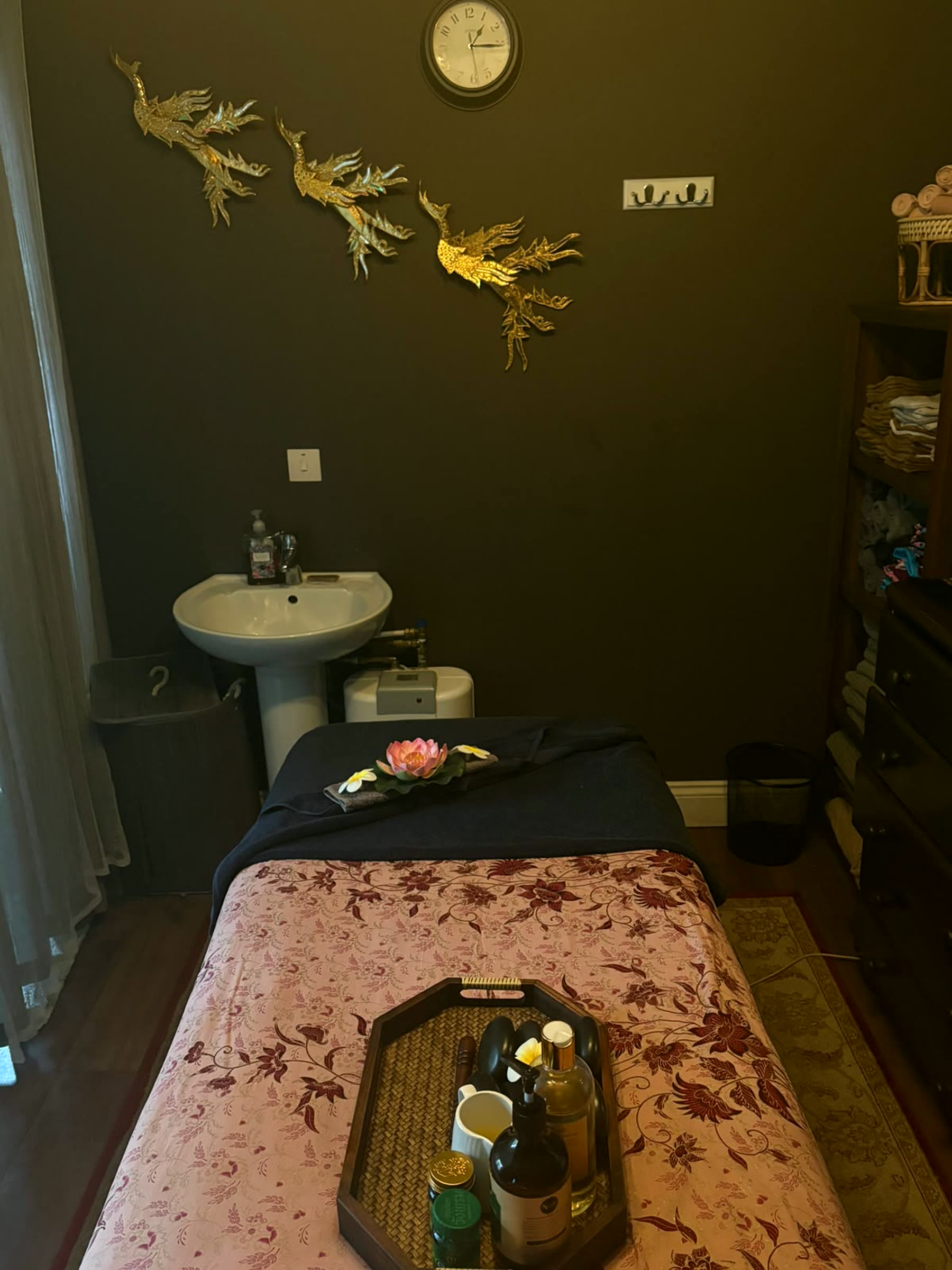 The treatment room's wash basin corner with gold phoenix wall art, a wall clock, and folded towels stored on a dresser